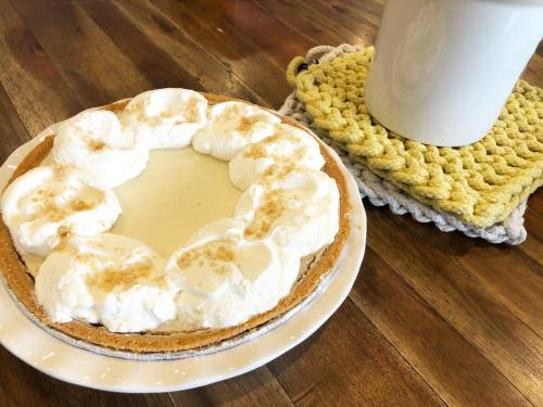 A creamy pie topped with whipped cream sits on a plate next to a white mug on a textured coaster.