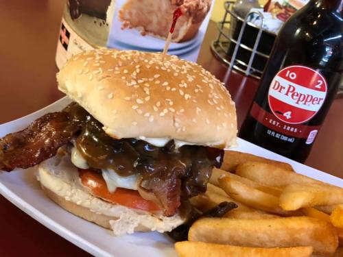 A sesame seed bun burger with bacon, cheese, and sauce, served with fries and a bottle of Dr Pepper.