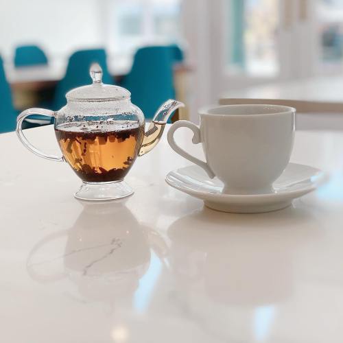 A glass teapot filled with tea beside a white cup on a saucer, set on a marble table.