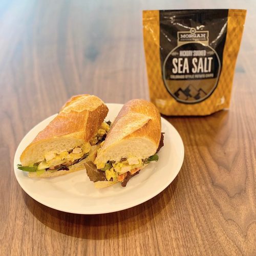 A sandwich with lettuce, egg, and vegetables on a plate, next to a bag of sea salt on a wooden table.