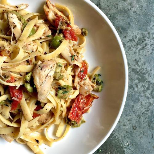 A plate of fettuccine pasta with chicken, sun-dried tomatoes, olives, and herbs on a textured surface.