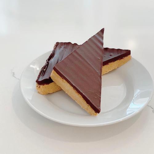 Two triangular chocolate-covered dessert bars on a white plate, resting on a light-colored surface.