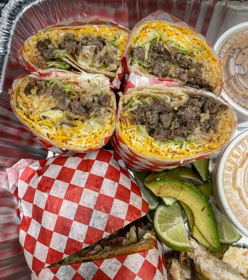 A tray of burritos with beef, cheese, and lettuce, accompanied by lime wedges and sauces in containers.