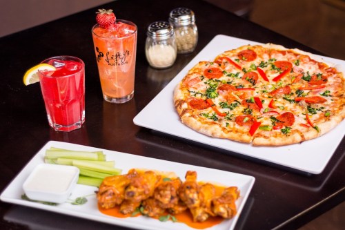 A table with a pepperoni pizza, chicken wings with sauce, and two colorful drinks.