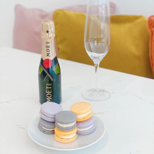 A mini bottle of Moët champagne next to a glass and a plate of colorful macarons on a white table.