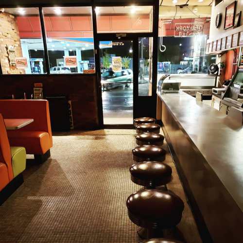A diner interior at night with colorful seating, a counter, and stools, viewed from the entrance. Rain is visible outside.