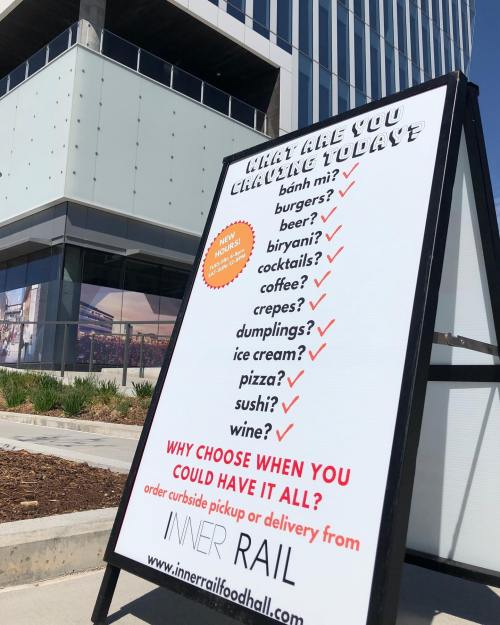 A sign outside a food hall lists various food options available for curbside pickup or delivery.