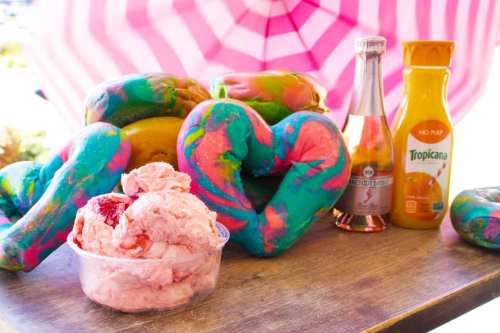 Colorful, swirled donuts and a bowl of pink ice cream, with drinks in the background, under a pink umbrella.