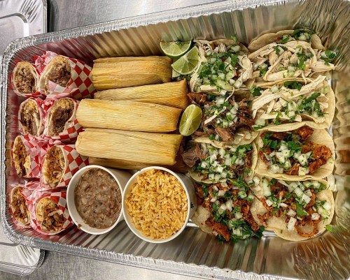 A tray of assorted Mexican food featuring tacos, tamales, rice, beans, and lime wedges.