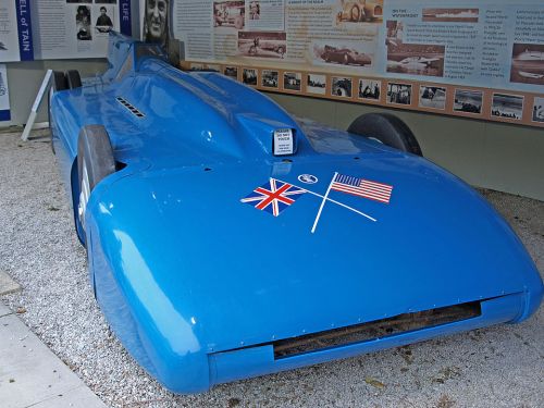 A blue racing car with British and American flags displayed, set against an informational backdrop.