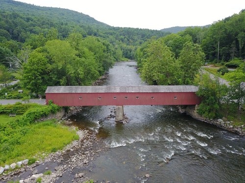 A red covered bridge spans a river, surrounded by lush green trees and rolling hills.