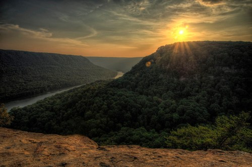 Sunset over a lush green valley with a winding river, framed by mountains under a dramatic sky.