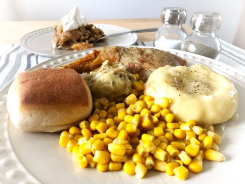 A plate of food featuring corn, mashed potatoes, stuffing, a roll, and fried chicken, with dessert in the background.