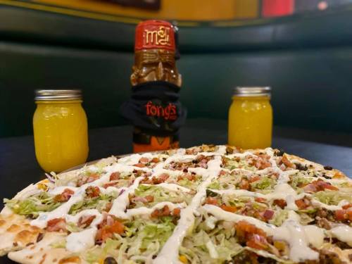 A pizza topped with lettuce, tomatoes, and sauce, flanked by two jars of yellow drink and a tiki mug in the background.