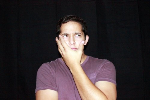 A young man with a puzzled expression rests his chin on his hand against a dark background.
