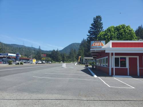 A quiet roadside scene featuring a cafe, empty parking lot, and mountains in the background under a clear blue sky.