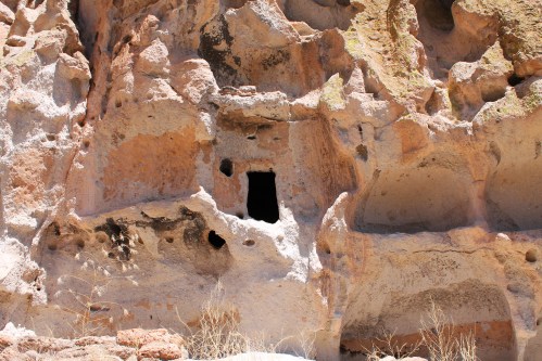 A rocky cliff face with a dark cave entrance and several small holes, surrounded by dry vegetation.