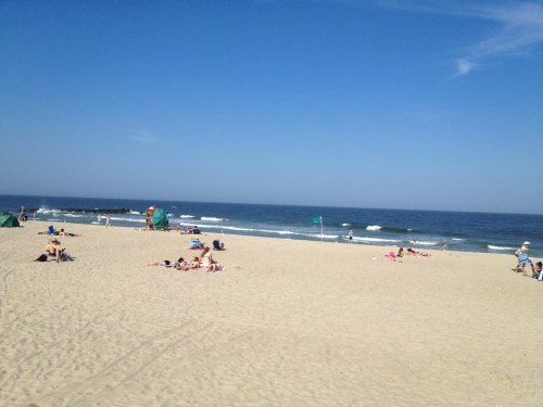 A sunny beach scene with people sunbathing, playing, and waves gently lapping at the shore under a clear blue sky.