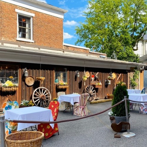Outdoor dining area with tables, rustic decor, and greenery against a brick wall and blue sky.