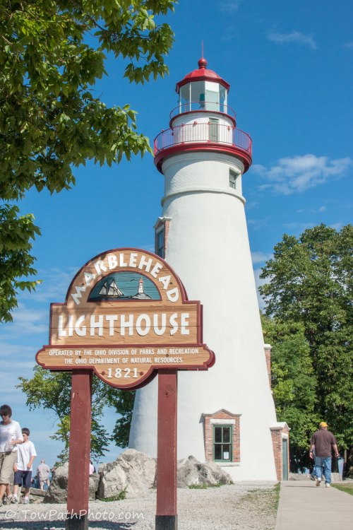 A tall white lighthouse with a red top, next to a sign reading "Marblehead Lighthouse," surrounded by trees and blue sky.