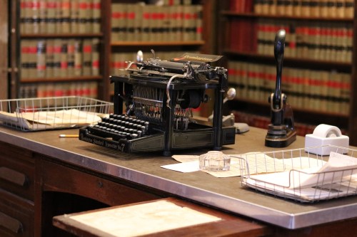 An antique typewriter on a wooden desk, surrounded by bookshelves filled with law books and scattered papers.