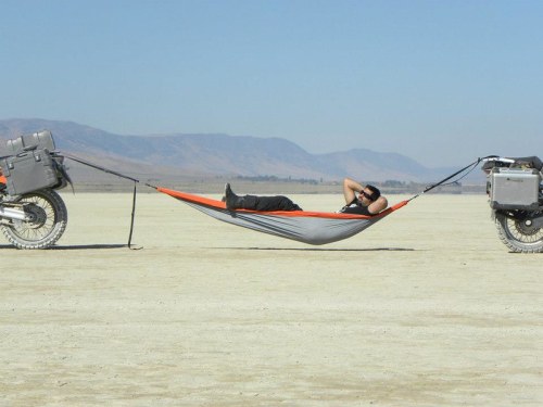 A person relaxes in a hammock suspended between two motorcycles on a dry, open landscape.