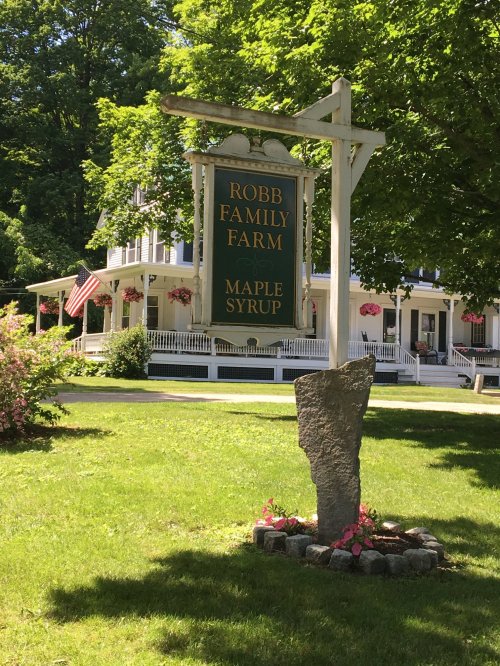 Sign for Robb Family Farm, featuring "Maple Syrup," with a charming house and greenery in the background.