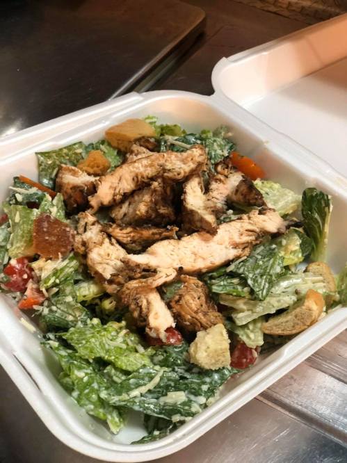 A takeout container filled with a Caesar salad topped with grilled chicken, croutons, and mixed greens.