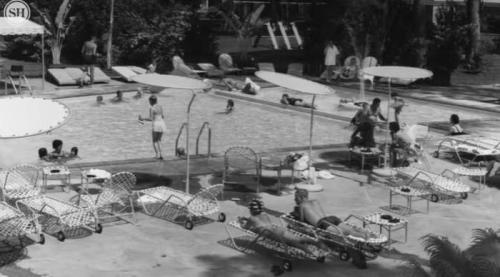A black-and-white image of a busy pool area with people swimming, lounging, and enjoying the sun.