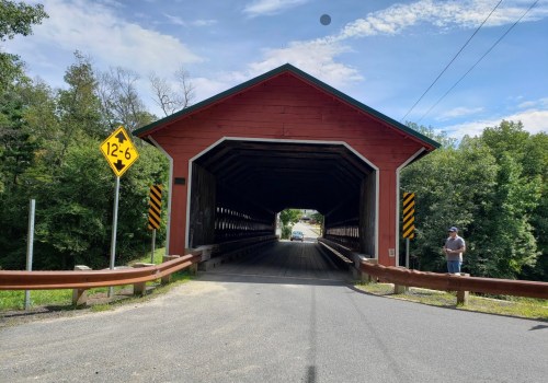 A red covered bridge with a wooden entrance, surrounded by trees, and a person standing nearby on a sunny day.