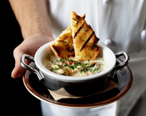 A chef holds a bowl of creamy soup topped with herbs and accompanied by a slice of grilled bread.