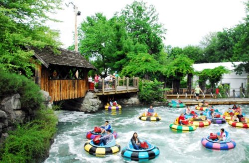 Colorful bumper boats float on a river, surrounded by lush greenery and a wooden bridge in the background.