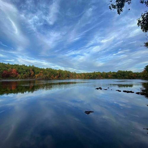 A serene lake reflects colorful autumn trees and a blue sky with wispy clouds.
