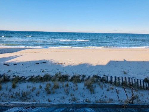 A serene beach scene with gentle waves, soft sand, and a wooden fence in the foreground under a clear blue sky.