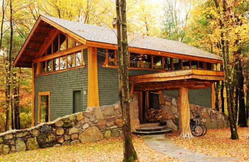 A modern cabin with wooden accents surrounded by autumn foliage and a stone pathway, featuring a bike by the entrance.