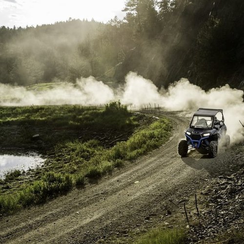 A blue ATV drives along a dusty trail near a serene lake, surrounded by trees and misty mountains.
