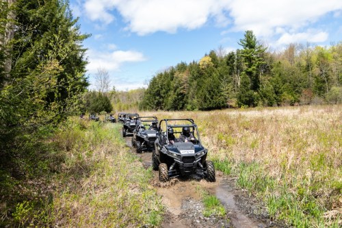 A line of off-road vehicles driving through a grassy, muddy trail surrounded by trees under a blue sky.
