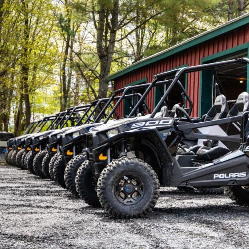 A row of black Polaris 900 off-road vehicles parked on gravel near a red barn surrounded by trees.