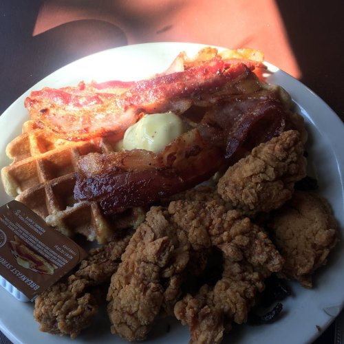 A plate of waffles topped with bacon, fried chicken, and a dollop of butter, accompanied by a syrup packet.