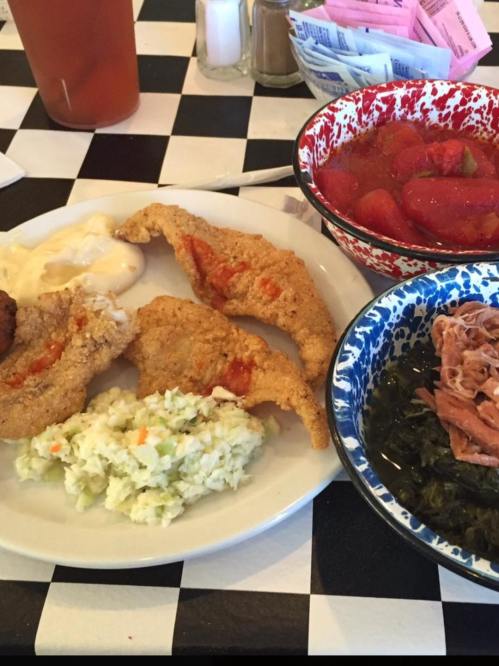A plate of fried fish fillets, coleslaw, and greens, accompanied by a bowl of stewed tomatoes.