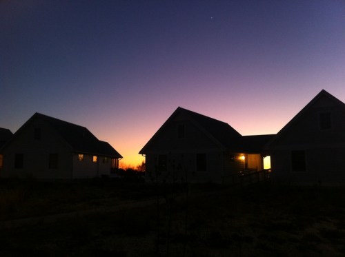 Silhouetted houses against a vibrant sunset sky, with warm lights glowing from the windows.