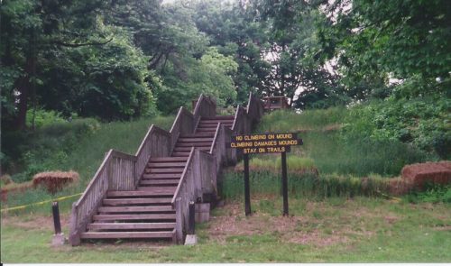 Wooden stairs lead up a grassy area, with a sign warning against climbing on the mound and to stay on trails.