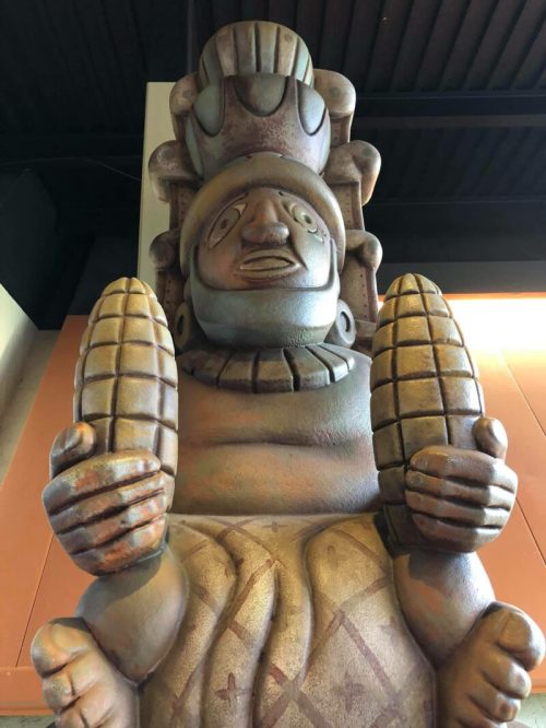 A large, stylized statue of a figure holding two corn cobs, with intricate details and a headdress.