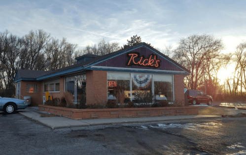 A cozy restaurant named "Rick's" with an open sign, surrounded by trees and a sunset in the background.