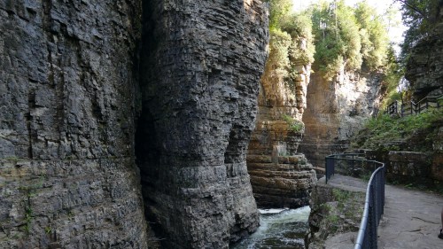 A narrow canyon with steep rock walls and a flowing river, surrounded by lush greenery and a pathway along the edge.