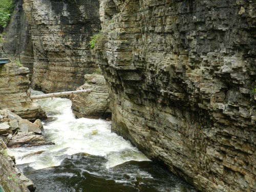 A rocky canyon with steep walls, featuring a flowing river and a fallen log across the water.