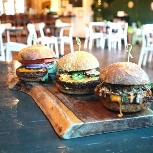 Three gourmet burgers on a wooden board, featuring various toppings, set in a cozy restaurant environment.