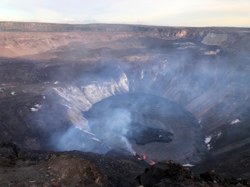 Aerial view of a volcanic crater with smoke and lava visible, surrounded by rugged terrain and distant mountains.