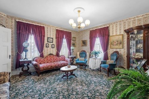 A vintage living room with floral wallpaper, ornate furniture, and elegant curtains, featuring a patterned carpet and decorative plants.