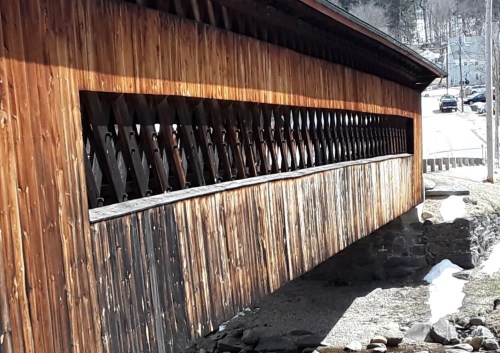 A wooden covered bridge with a slatted design, spanning over a rocky area, surrounded by snow and trees.